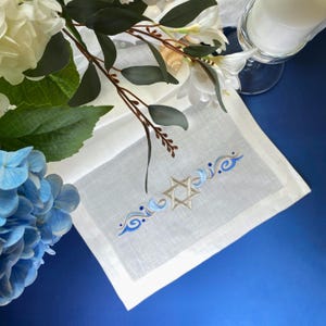 May include: White linen napkin with an embroidered Star of David in the center, flanked by blue and silver wave designs. The napkin is surrounded by white and blue flowers, and a candle in a glass holder.