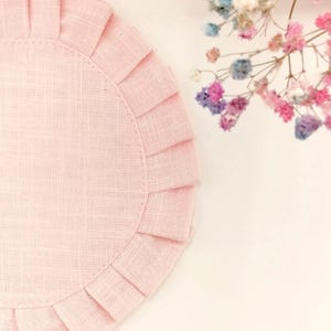 Op de afbeelding: Een lichtroze, ronde placemat met een gerimpelde rand. De placemat is gemaakt van een textiel met textuur en staat tegen een witte achtergrond. Kleurrijke droogbloemen zijn op de achtergrond.