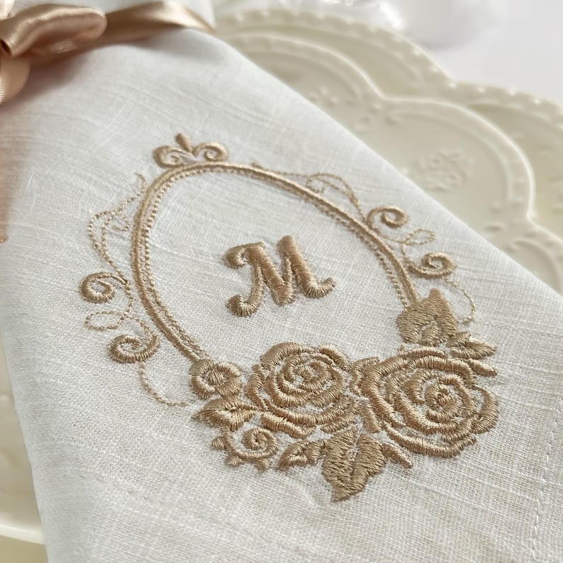 Puede incluir: Una servilleta de tela blanca con un bordado de color dorado de la letra M centrada dentro de un marco ovalado. El dise&ntilde;o incluye motivos florales de rosas en la parte inferior del marco y una cinta dorada clara atada en la esquina superior.