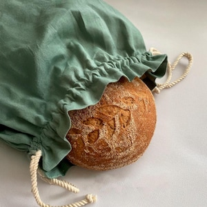 May include: A green linen bread bag with a drawstring closure. The bag is filled with a loaf of sourdough bread with a decorative pattern on top.