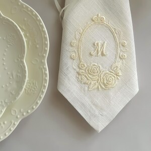 Puede incluir: Una servilleta de lino color crema con un marco ovalado bordado, una letra M central y motivos florales de rosas. Junto a la servilleta hay dos platos color crema con bordes festoneados y patrones florales en relieve en los bordes.