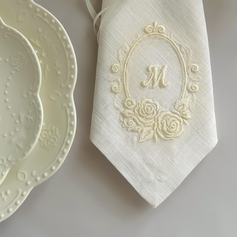 Puede incluir: Una servilleta de lino color crema con un marco ovalado bordado, una letra M central y motivos florales de rosas. Junto a la servilleta hay dos platos color crema con bordes festoneados y patrones florales en relieve en los bordes.
