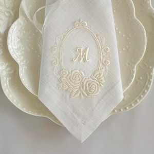 Puede incluir: Una servilleta de lino blanco con un marco ovalado bordado en color blanco hueso, con volutas decorativas y motivos florales de rosas en la base. El centro del &oacute;valo contiene la letra M bordada en una fuente cursiva centrada en la tela.