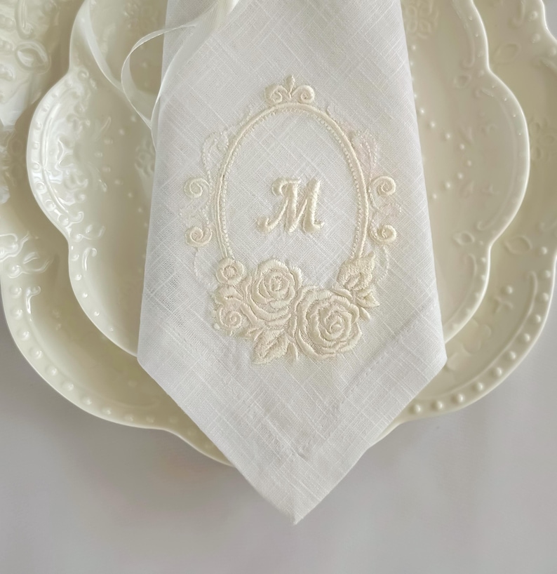 Puede incluir: Una servilleta de lino blanco con un marco ovalado bordado en color blanco hueso, con volutas decorativas y motivos florales de rosas en la base. El centro del &oacute;valo contiene la letra M bordada en una fuente cursiva centrada en la tela.