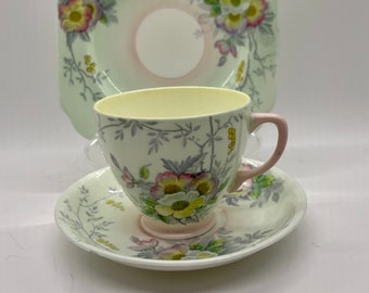 May include: A vintage teacup and saucer set with a floral design. The cup and saucer are white with a green and pink floral pattern. The cup has a handle and the saucer is round.