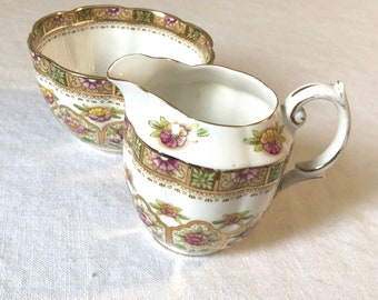 May include: A vintage cream-colored sugar bowl and creamer set with a floral pattern in shades of pink, yellow, and green. The set has a gold trim and a scalloped edge.