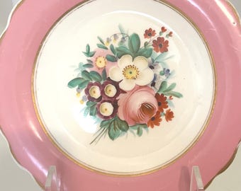 May include: A white porcelain plate with a pink rim and gold trim. The center of the plate features a hand-painted bouquet of pink, white, and red flowers.