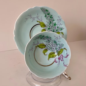 Könnte beinhalten: Ein hellblaues Tee-Set mit Blumenmuster. Tasse und Untertasse sind mit einem Strauß violetter und weißer Blumen mit grünen Blättern verziert. Die Blumen sind mit einem rosa Band akzentuiert.