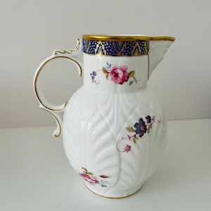 May include: A white porcelain pitcher featuring a textured leaf pattern, a gold-trimmed rim, and a dark blue band with gold accents. The body is decorated with hand-painted floral motifs in pink, purple, and green, complemented by a gold-edged handle.