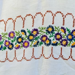 May include: A white fabric featuring a central horizontal band of embroidered flowers in shades of blue, purple, and yellow with green leaves. The floral design is bordered above and below by a scalloped pattern stitched in brown thread.