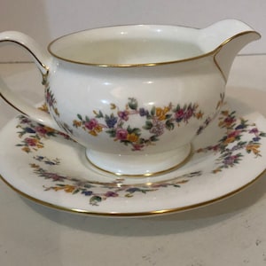 May include: A white porcelain gravy boat with a floral pattern and gold trim. The boat is sitting on a matching saucer.