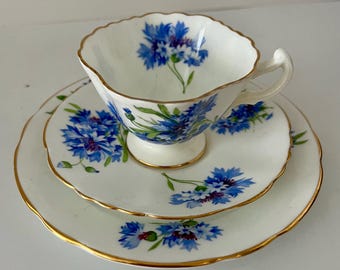 May include: A vintage tea set comprising a teacup, saucer, and side plate. Each piece is white with a blue floral design and gold edging. The teacup has a scalloped rim and a pedestal base.
