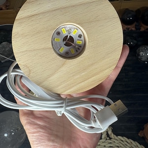 Puede incluir: Una base de madera redonda y clara con un área circular empotrada que alberga una pequeña lámpara LED. Un cable USB blanco está conectado. La base se sostiene en una mano, con otros objetos en el fondo.