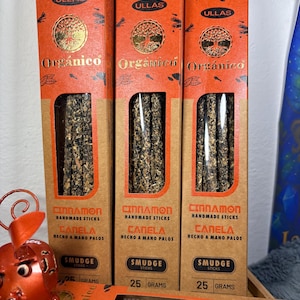 Könnte beinhalten: Drei Schachteln Zimt-Smudge-Sticks. Die Schachteln sind orange und braun mit dem Wort "ULLAS" oben. Auf den Schachteln stehen die Wörter "Organico", "Cinnamon Handmade Sticks" und "Canela Hecho a Mano Palos". Jede Schachtel enthält 25 Gramm Produkt.