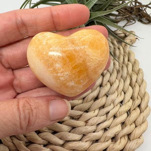 Può includere: Una pietra lucidata a forma di cuore, nei toni dell'arancione e del crema. La pietra è tenuta in mano, con un sottobicchiere intrecciato e una pianta verde sullo sfondo. La pietra ha una finitura liscia e lucida.