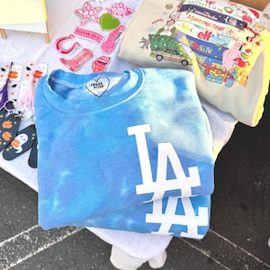 Puede incluir: Una sudadera azul teñida con el logo blanco de LA. La sudadera está doblada sobre una superficie blanca, con otros artículos como pegatinas y una sudadera con temática navideña visibles en el fondo. También hay llaveros con temática de Halloween.