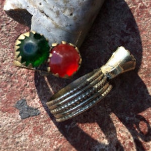 May include: A silver ring with three stones, one green, one red, and one clear. The ring is on a textured surface.
