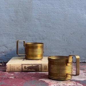 May include: Two brass-colored metal mugs with rectangular handles. The mugs have a ribbed design and sit on a vintage book. The background is a weathered blue wall. The mugs are likely vintage or antique.