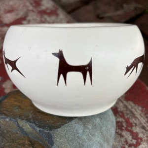 May include: A white ceramic bowl with a rounded shape. The bowl features a repeating pattern of stylized brown dog silhouettes around its exterior. The bowl is sitting on a grey rock.
