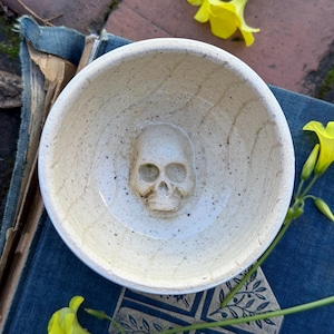 May include: A cream-coloured ceramic bowl featuring a skull-shaped relief in the centre. The bowl has a speckled texture and a shallow depth. The skull is detailed with eye sockets, a nose cavity, and teeth. The bowl is likely handmade and suitable for decorative use.