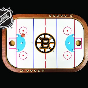 May include: A wooden tabletop hockey game with a white playing surface, blue and red markings, and a brown puck. The game features the Boston Bruins logo in the center of the playing surface. The game is surrounded by a wooden frame with brass accents.