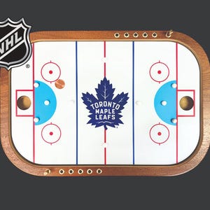Peut inclure: Un jeu de hockey de table avec un cadre en bois et une surface de jeu blanche. Le jeu présente le logo des Maple Leafs de Toronto et le logo de la LNH. Le jeu comprend une rondelle et deux filets.