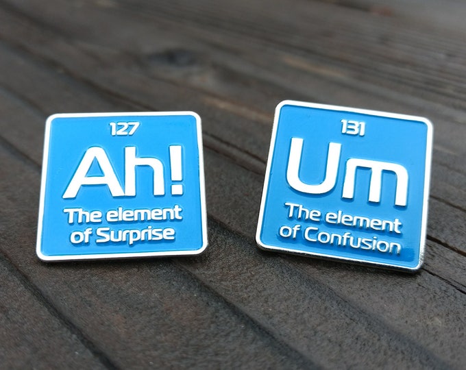 May include: Two blue enamel pins with white lettering. The first pin says "Ah! The element of Surprise" with the number 127 above it. The second pin says "Um The element of Confusion" with the number 131 above it.