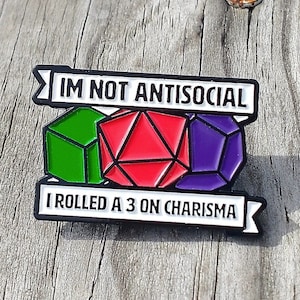 Op de afbeelding: Een zwart-witte emaille pin met een wit banner waarop staat "Ik ben niet antisociaal, ik heb een 3 gegooid op charisma". De banner wordt geflankeerd door drie dobbelstenen, een groene, een rode en een paarse.