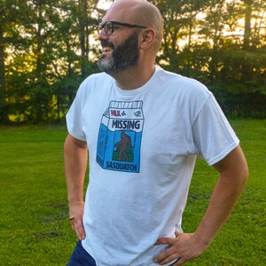 May include: A white t-shirt with a blue and white graphic of a milk carton with the text "Milk & Missing Sasquatch".
