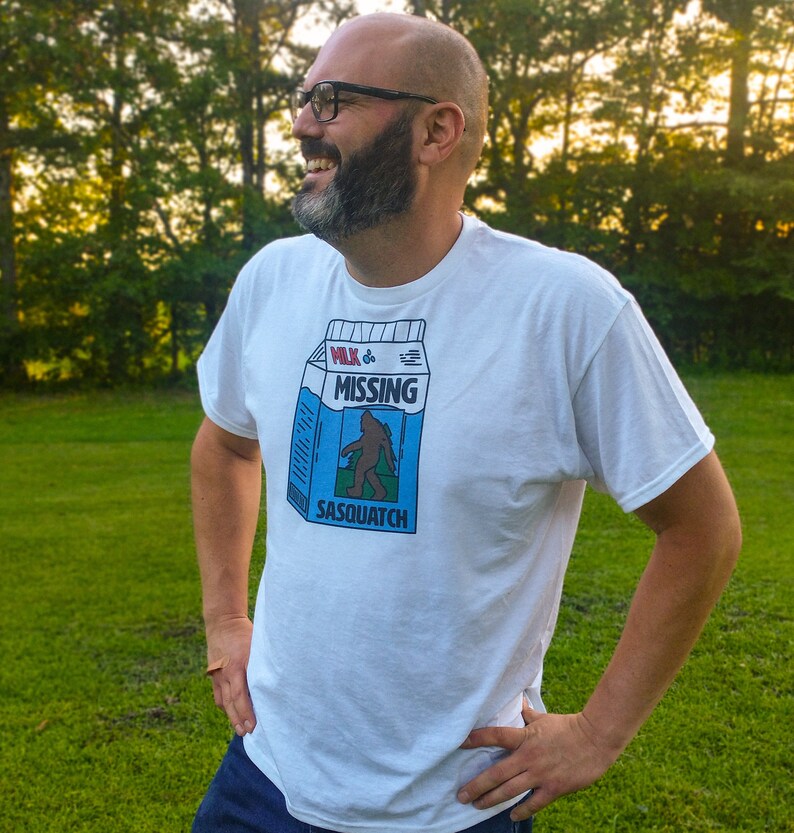 May include: A white t-shirt with a blue and white graphic of a milk carton with the text "Milk & Missing Sasquatch".
