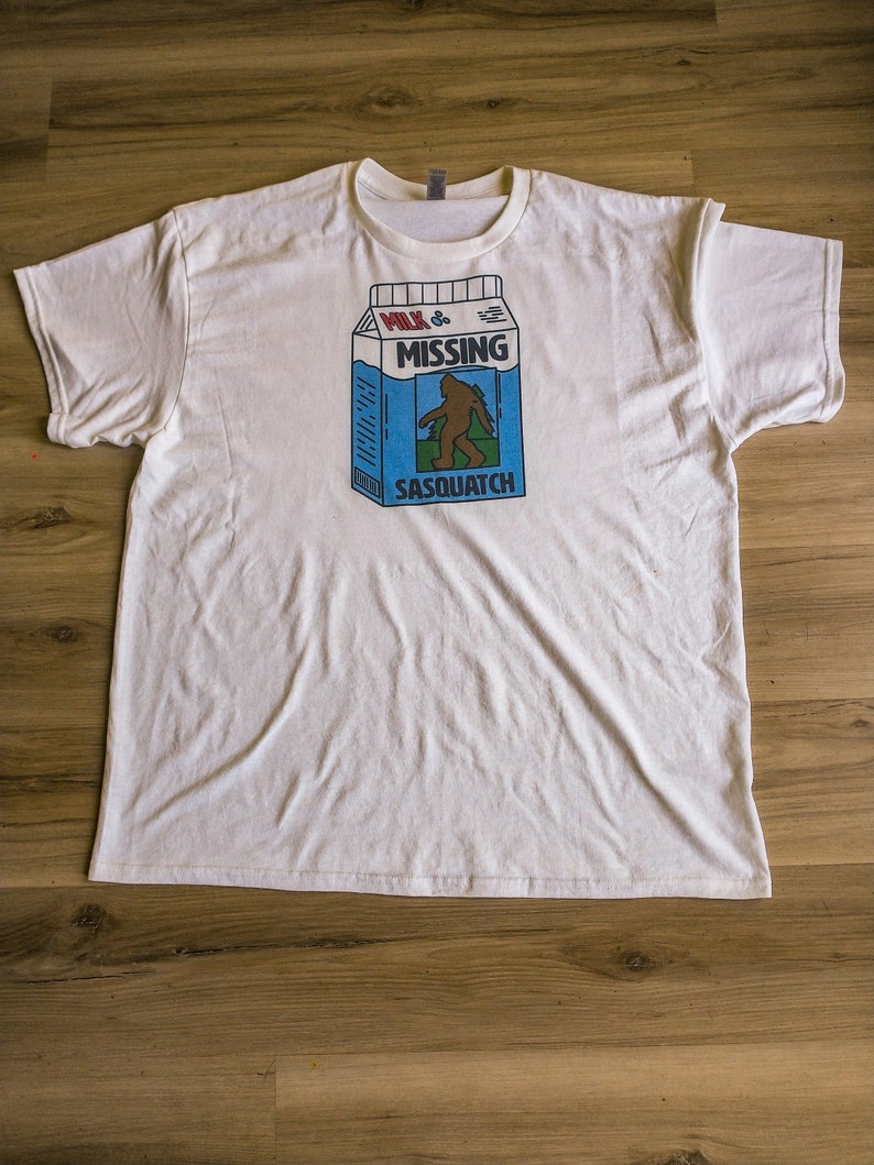 May include: White t-shirt with a graphic of a milk carton that says "Milk is Missing Sasquatch".