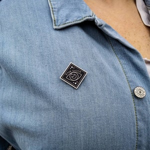May include: A black enamel pin with a white outline of an eye with a crescent moon inside. The pin is shaped like a diamond and says "Star Gazer" around the eye.