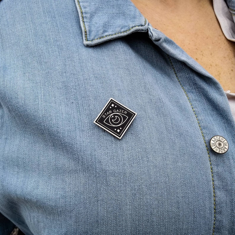 May include: A black enamel pin with a white outline of an eye with a crescent moon inside. The pin is shaped like a diamond and says "Star Gazer" around the eye.