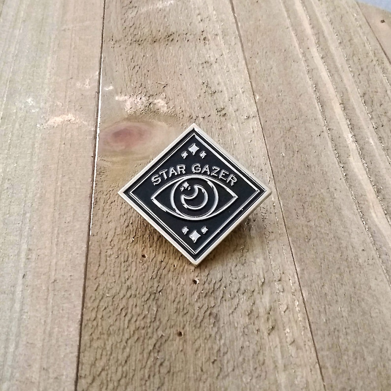 May include: Black and silver enamel pin with a diamond shape. The pin features a stylized eye with a crescent moon inside and the text "STAR GAZER" around the eye.