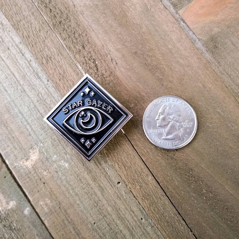 May include: A black and silver enamel pin with a diamond shape. The pin features a stylized eye with a crescent moon inside and the text "STAR GAZER" around the eye. The pin is about the size of a US quarter.