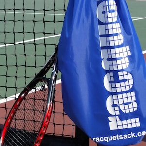 Puede incluir: Una bolsa de raqueta de tenis azul con el texto "racketsack.co" y la palabra "sack" impresa en ella. Tres pelotas de tenis amarillas están en una cancha de tenis de arcilla roja.
