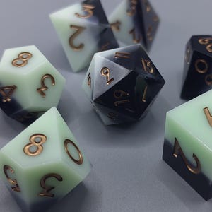 Puede incluir: Juego de siete dados poliédricos bicolores. Los dados presentan un diseño de dos tonos, con una combinación de colores verde claro y negro. Los números son de color cobre. Estos dados son para juegos de mesa.