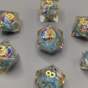 May include: Set of seven clear resin dice with gold numbering and colorful swirling designs. The dice have a blue glitter background and are arranged on a gray surface. The dice are for role-playing games.