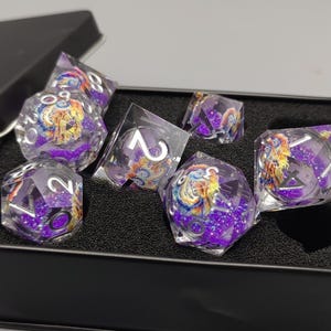 May include: A set of seven polyhedral dice in a black metal box. The dice are clear with purple glitter and feature colorful artwork. The dice are for role-playing games.