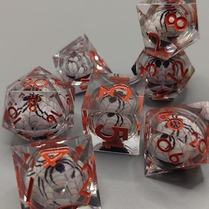 May include: Set of seven clear dice with a spider and web design inside. The numbers are in orange. The dice are for role-playing games.
