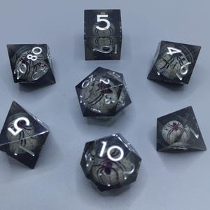 Peut inclure: Un ensemble de sept dés polyédriques en résine sombre transparente, contenant chacun une petite sphère blanche avec un motif d'araignée noire et rouge et une toile. Les chiffres sont gravés en argent sur les faces extérieures des dés.