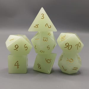 Peut inclure: Un ensemble de six dés lumineux de différentes formes géométriques, dont une pyramide, un cube et plusieurs polyèdres. Les dés sont vert pâle avec des chiffres dorés, parfaits pour les jeux de rôle.