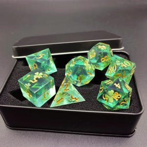 K&ouml;nnte beinhalten: Ein Satz von sieben polyedrischen W&uuml;rfeln in einer schwarzen Metallbox. Die W&uuml;rfel sind transparent gr&uuml;n mit goldenen Zahlen und Kleemotiven. Die W&uuml;rfel sind f&uuml;r Rollenspiele.