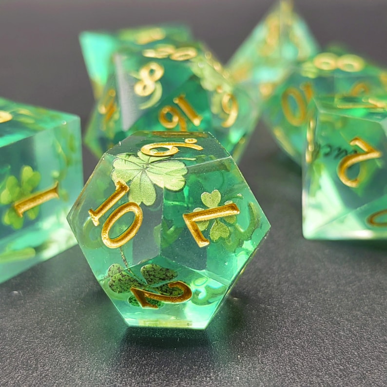 K&ouml;nnte beinhalten: Satz transluzenter gr&uuml;ner polyedrischer W&uuml;rfel mit goldenen Zahlen und Kleeblatt-Designs. Die W&uuml;rfel f&uuml;r Tischspiele haben verschiedene Formen, darunter ein 20-seitiger W&uuml;rfel. Die W&uuml;rfel liegen auf einer dunklen Oberfl&auml;che.