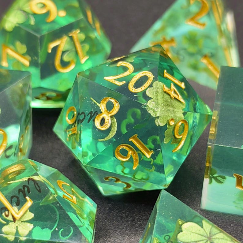 K&ouml;nnte beinhalten: Ein Satz transluzenter gr&uuml;ner polyedrischer W&uuml;rfel mit goldfarbenen Zahlen und Kleeblattmustern. Die W&uuml;rfel sind f&uuml;r Tabletop-Rollenspiele konzipiert. Der zwanzigseitige W&uuml;rfel ist prominent dargestellt, andere W&uuml;rfel sind teilweise sichtbar.