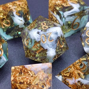Peut inclure: Gros plan sur un ensemble de dés polyédriques. Les dés sont transparents avec des chiffres et des inclusions dorés, et un mélange de tourbillons blancs et bleu-vert. Les dés sont disposés sur une surface sombre.