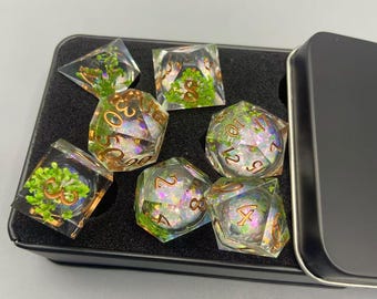 May include: Set of seven clear resin dice with gold numbering and green plant inclusions, presented in a black metal tin. The dice are various geometric shapes, including a d4, d6, d8, d10, d12, d20, and a d30.
