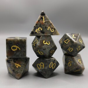 May include: Set of seven polyhedral dice in shades of gray, brown, and black with gold-colored numbering. The dice are made of a speckled stone-like material. The set includes a d4, d6, d8, d10, d12, d20, and a percentile die.