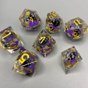Peut inclure: Ensemble de sept dés en résine transparente, avec des chiffres dorés et un motif de paillettes violettes et de chat noir. Les dés sont destinés aux jeux de société. Chaque dé a une forme et un nombre de faces différents.