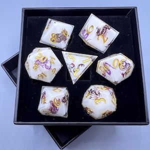May include: A set of seven polyhedral dice in a black box. The dice are white with gold numbering and purple and brown dragon illustrations. The dice include a D4, D6, D8, D10, D12, D20, and a triangular D3.